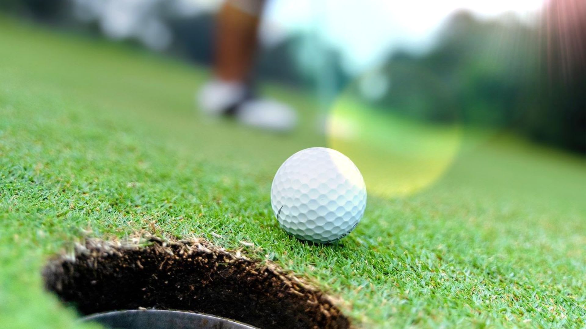 Close-up of a golf ball near the edge of a hole on a green field, with a golfer's legs visible in the blurred background.
